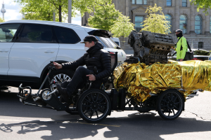 Ein Mann im Rollstuhl mit einem großen Motor auf dem Rücken, umgeben von Fahrzeugen auf einer Straße mit Bäumen, Gebäuden, Polen und einem klaren blauen Himmel im Hintergrund; er trägt eine schwarze Jacke, eine Mütze und hält ein Objekt in der Hand.