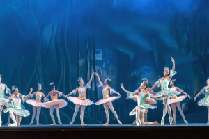 Eine Gruppe von Balletttänzern in farbenfrohen Kostümen, die *Der Nussknacker* auf der Bühne des Royal Opera House tanzen, bewegen sich synchronisiert anmutig gegen einen blauen Hintergrund.