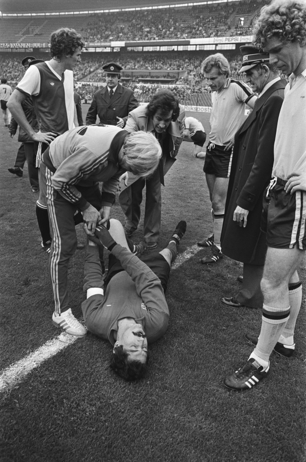 Schwarzes und weiße Bild eines Förspielers, der auf dem Boden liegt und von einer Gruppe von Mönnen umgeben ist, mit einer Menge im Stadionhintergrund.