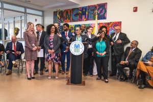 Gruppe von Menschen vor einem Rednerpult mit Mikrofon und einer Tafel mit der Aufschrift "Sancho Stadtrat tritt dem Stadtrat bei", mit sitzenden und stehenden Teilnehmern, Flaggen, einem Feuerlöscher und Deckenleuchten im Hintergrund.