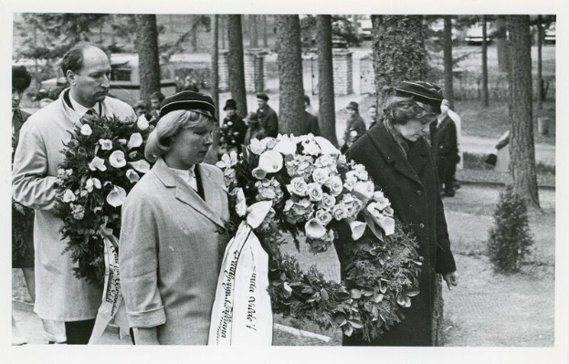 Schwarzes und weißes Foto einer Gruppe von Menschen, die Blumensträuße bei einer Gedenkfeier für Holocaust-Opfer halten, mit Bäumen und Gebäuden im Hintergrund.