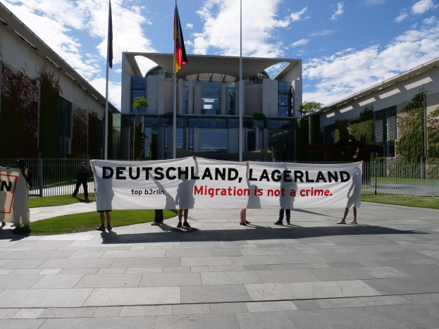 Gruppe von Menschen mit einem Banner mit der Aufschrift "Deutschland, Lagerland Migration ist kein Verbrechen" vor einem Gebäude mit Fenstern, umgeben von Gras, einem Metallzaun, Pflanzen, Bäumen und Fahnenmasten unter einem bewölkten Himmel.