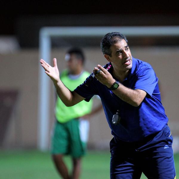Ein Mann in einem blauen T-Shirt und Hose steht auf einem Fußballfeld und hält ein Mikrofon in der Hand, während er als Trainer gestikuliert. Im Hintergrund sind ein Torpfosten, eine weitere Person in einem grünen Kleid und eine Wand zu sehen.