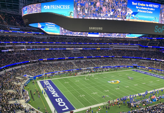 Ein großes Stadion mit Zuschauern, die ein Fußballspiel auf grünem Rasen verfolgen, mit Spielern auf dem Feld und einem großen Bildschirm mit Text und Bildern oben.