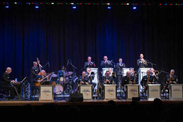 Eine Gruppe Musiker spielt auf einer hell erleuchteten Bühne mit Mikrofonen, Ständern und Texttafeln, die 'The New Orleans Jazz Band' lesen, während das Publikum von unten zusieht.