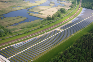 Eine Luftaufnahme eines großen Solarparks mit Solarpanelen in einem Feld, umgeben von Bäumen, Gras, Pflanzen und Wasser, mit einem Zug, der auf einem Bahngleis fährt.
