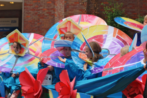 Eine Gruppe von Kindern in bunten Kostümen fährt auf einem Festwagen mit Schirmen, mit einem Gebäude, Pflanzen und Lichtern im Hintergrund.