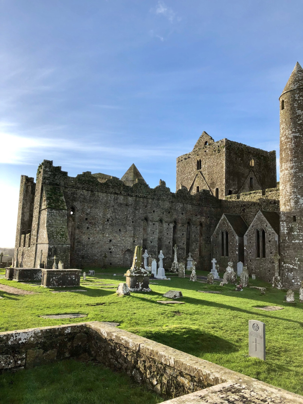Ruinen einer alten irischen Abtei mit einem Friedhof im Vordergrund und einer Burg im Hintergrund bei bewölktem Himmel.