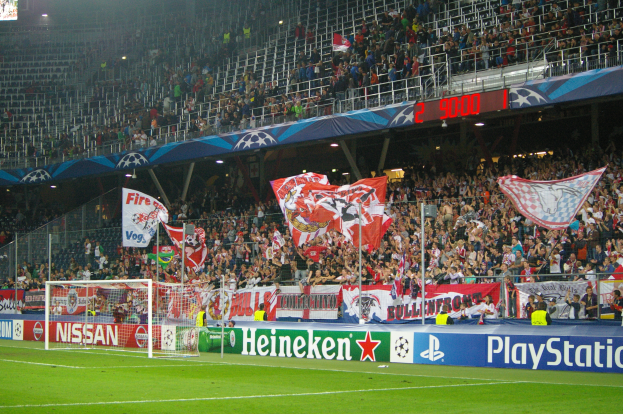 Ein Fussballspiel wird in einem gut beleuchteten Stadion mit einer grossen Zuschauermenge, grünem Rasen, einem mit Bannern geschmückten Torpfosten und einem Bildschirm in der oberen linken Ecke gespielt.