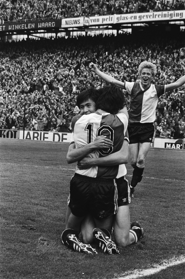 Drei Fussballspieler umarmen sich auf dem Boden nach einem Tor, mit jubelnder Menge und schwenkenden Fahnen im Hintergrund, auf einem Schwarz-Weiss-Bild.