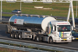 Großer Tanklastwagen fährt auf einer Autobahn neben einem Auto, mit sichtbaren Insassen im Truck und Gras, Stangen und anderen Gegenständen im Hintergrund.