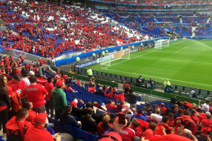 Eine große Menschenmenge in einem Stadion schaut einem Fußballspiel zu, mit Menschen auf Sitzen und Stehplätzen, Bannern, einem Torpfosten und Deckenbeleuchtung.