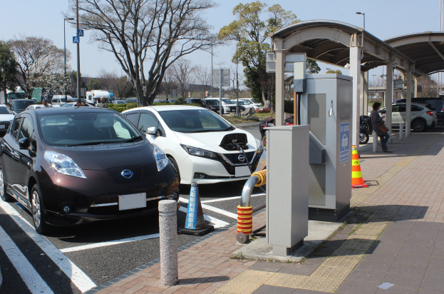 Elektroauto-Ladestation in Japan mit Autos auf der Straße, Verkehrskegel, einer Person auf dem Gehweg, einem Schuppen, Polen, Lampen, Schildern, Bäumen, Pflanzen und einem Himmel im Hintergrund.