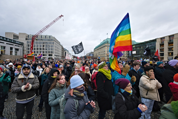 Große Gruppe von Menschen bei der LGBTQ+-Rechtsdemo in Berlin, mit Fahnen und Plakaten, vor einem Gebäude mit einem Kran und einem bewölkten Himmel.
