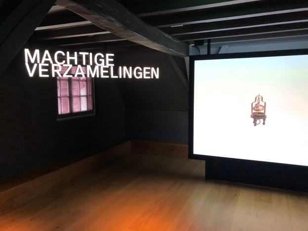 Installationsansicht der Ausstellung 'Machtige Verzamelingen' im Museum of Modern Art in Berlin, mit einem großen Bildschirm mit Text an der Wand, einem Fenster links und sichtbarem Decke.