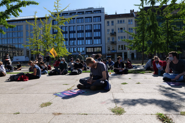 Eine Gruppe von Menschen sitzt vor einem Gebäude auf dem Boden während einer Demonstration in Berlin, einige tragen Masken, mit verstreuten Taschen und Gegenständen, unter einem klaren blauen Himmel und umgeben von Bäumen.