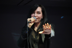 Eine Frau in einem schwarzen Kleid mit einem Dutt singt leidenschaftlich in ein Mikrofon auf einer hell erleuchteten Bühne, die Augen geschlossen und der Gesichtsausdruck intensiv.