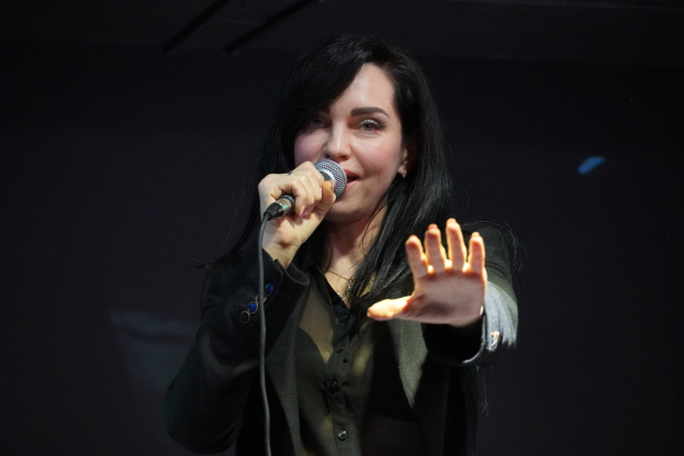 Eine Frau in einem schwarzen Kleid mit einem Dutt singt leidenschaftlich in ein Mikrofon auf einer hell erleuchteten Bühne, die Augen geschlossen und der Gesichtsausdruck intensiv.