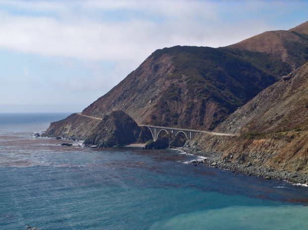 Eine Betonbogenbrücke, Bixby Creek Bridge, überspannt einen Wasserlauf mit Hügeln und Wolken im Hintergrund.