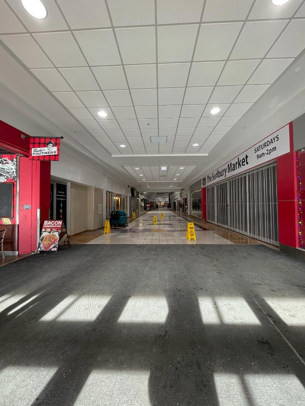 Innenansicht eines Einkaufszentrums mit Deckenbeleuchtung, einem Stuhl und einer Tafel, die links mit "Sydbury Market - Vorher und Nachher" beschriftet ist, sowie verstreuten Gegenständen rechts und auf dem Boden.