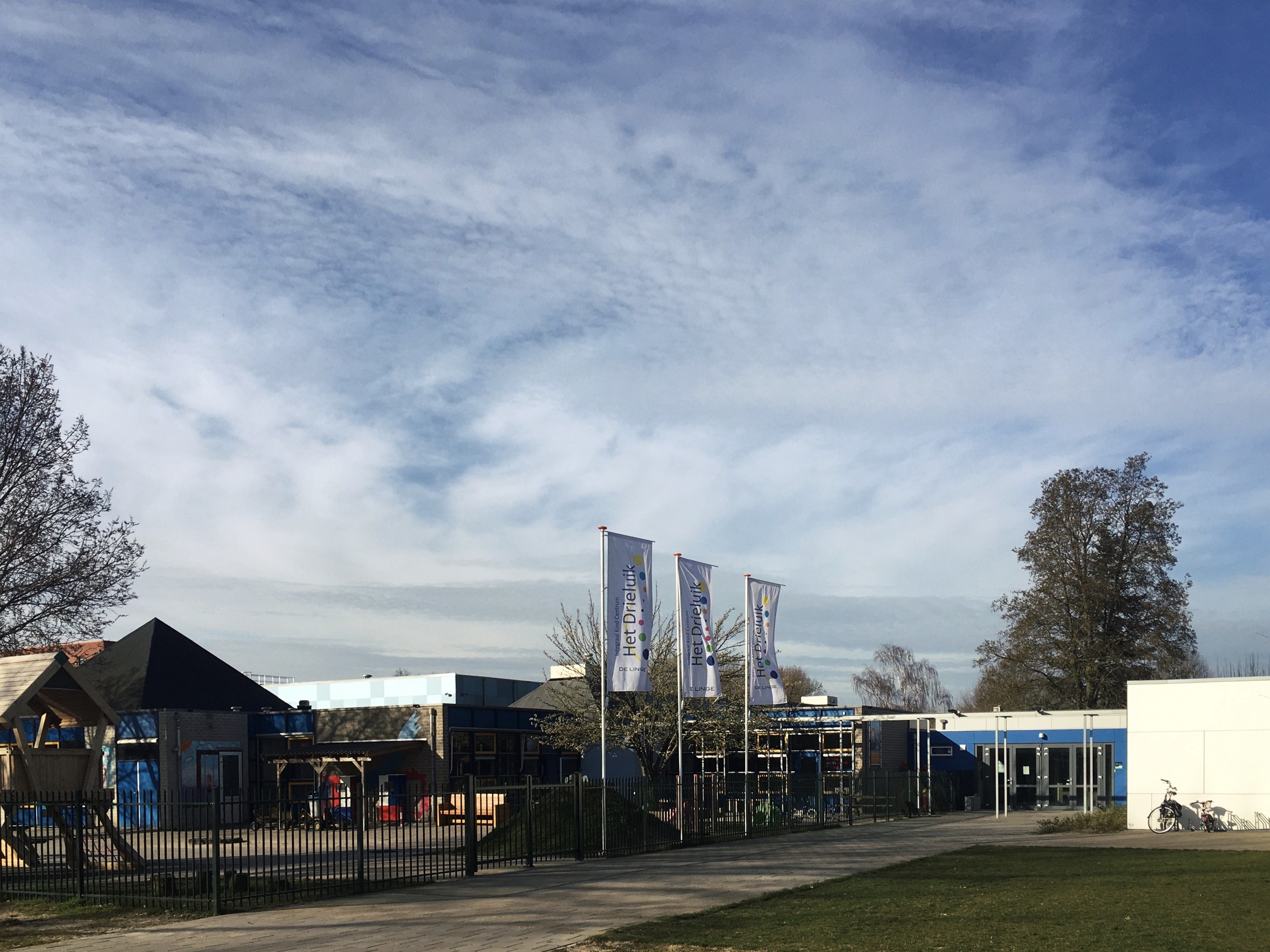 Großes Gebäude mit einem grasbewachsenen Bereich davor, umgeben von einem Metallzaun, mit einem Weg, Bäumen, Bannern, einem geparkten Fahrrad, nahegelegenen Häusern und einem bewölkten Himmel, identifiziert als der neue Spielplatz der Schule.