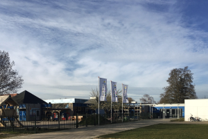Großes Gebäude mit einem grasbewachsenen Bereich davor, umgeben von einem Metallzaun, mit einem Weg, Bäumen, Bannern, einem geparkten Fahrrad, nahegelegenen Häusern und einem bewölkten Himmel, identifiziert als der neue Spielplatz der Schule.