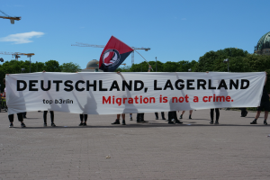 Eine Gruppe von Menschen hält eine Fahne mit der Aufschrift "Deutschland, Lagerland Migration ist kein Verbrechen" und eine Flagge, die auf dem Boden steht mit Straßenlaternen, Straßenlaternen, Bäumen, Kränen, einem Gebäude und einem bewölkten Himmel im Hintergrund.