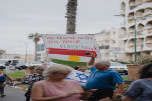 Eine Gruppe von Menschen marschiert nachts eine Straße entlang, hölt ein Schild mit der Aufschrift "Wir stehen zu den tapferen kurdischen Menschen" in der Hand, mit einem Van, der an der Straße geparkt ist, Straßenlaternen, Bäumen, Gebäuden und einem klaren blauen Himmel im Hintergrund.