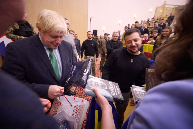 Ein Mann im Anzug und Krawatte signiert Autogramme für eine Menschenmenge, einige halten Taschen und Bücher, mit einer beleuchteten Wand im Hintergrund.