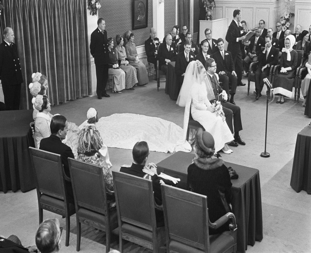Schwarzes und weißes Foto einer Hochzeitszeremonie mit sitzenden Gästen, einem Mann, der in ein Mikrofon spricht, einer Frau mit einem Blumenstrauß, einem Podium mit einem Mikrofon, Blumenschmuck, einem Vorhang, einem gerahmten Foto an der Wand und einer Tür im Hintergrund.