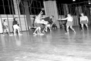 Eine Gruppe von Männern, die Handball auf einem Holzfußboden mit einer Wand und Lampen im Hintergrund spielen, in Schwarz-Weiß dargestellt.