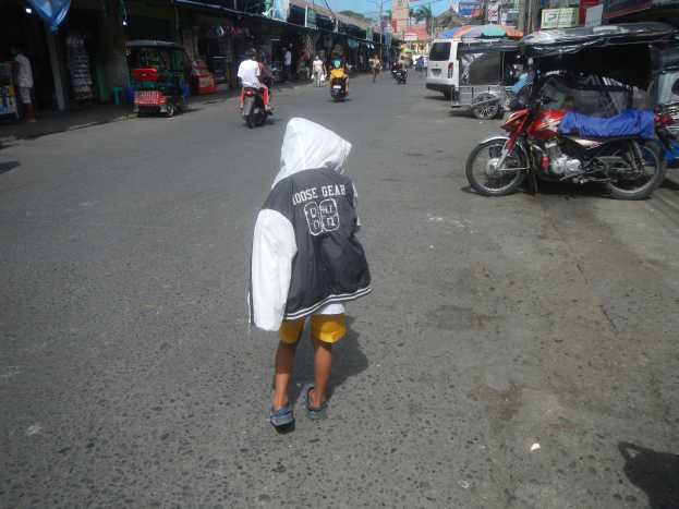 Ein junger Junge geht eine Straße entlang, die voller Motorräder, andere Fahrzeuge und Fußgänger ist, mit Gebäuden, Namensschildern, Bäumen, Pfählen und einem klaren blauen Himmel im Hintergrund.