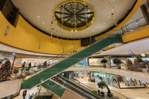 Ein belebtes Einkaufszentrum mit Rolltreppen, Geländern, Topfpflanzen, Geschäften, Texttafeln, Deckenleuchten und hängenden Dekorationen, mit einigen Menschen.