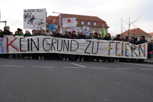 Demonstranten mit einem Banner mit der Aufschrift 'Kein Grund zur Feier' gegen deutsche Sparmaßnahmen, mit Straßeninfrastruktur und Gebäuden im Hintergrund bei klarem Himmel.
