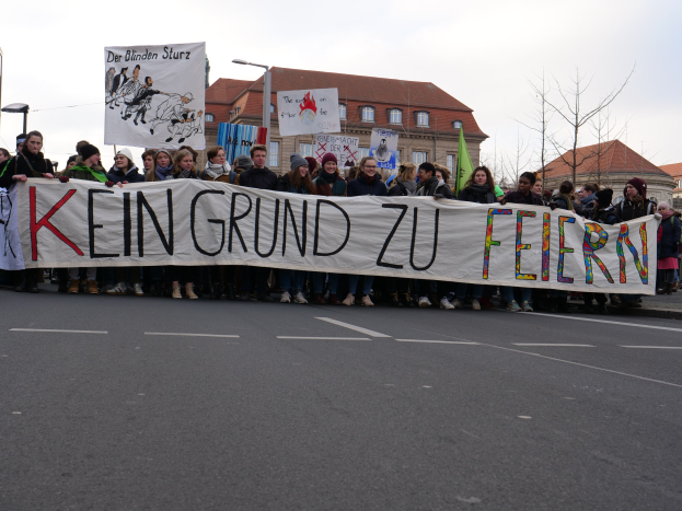 Demonstranten mit einem Banner mit der Aufschrift 'Kein Grund zur Feier' gegen deutsche Sparmaßnahmen, mit Straßeninfrastruktur und Gebäuden im Hintergrund bei klarem Himmel.