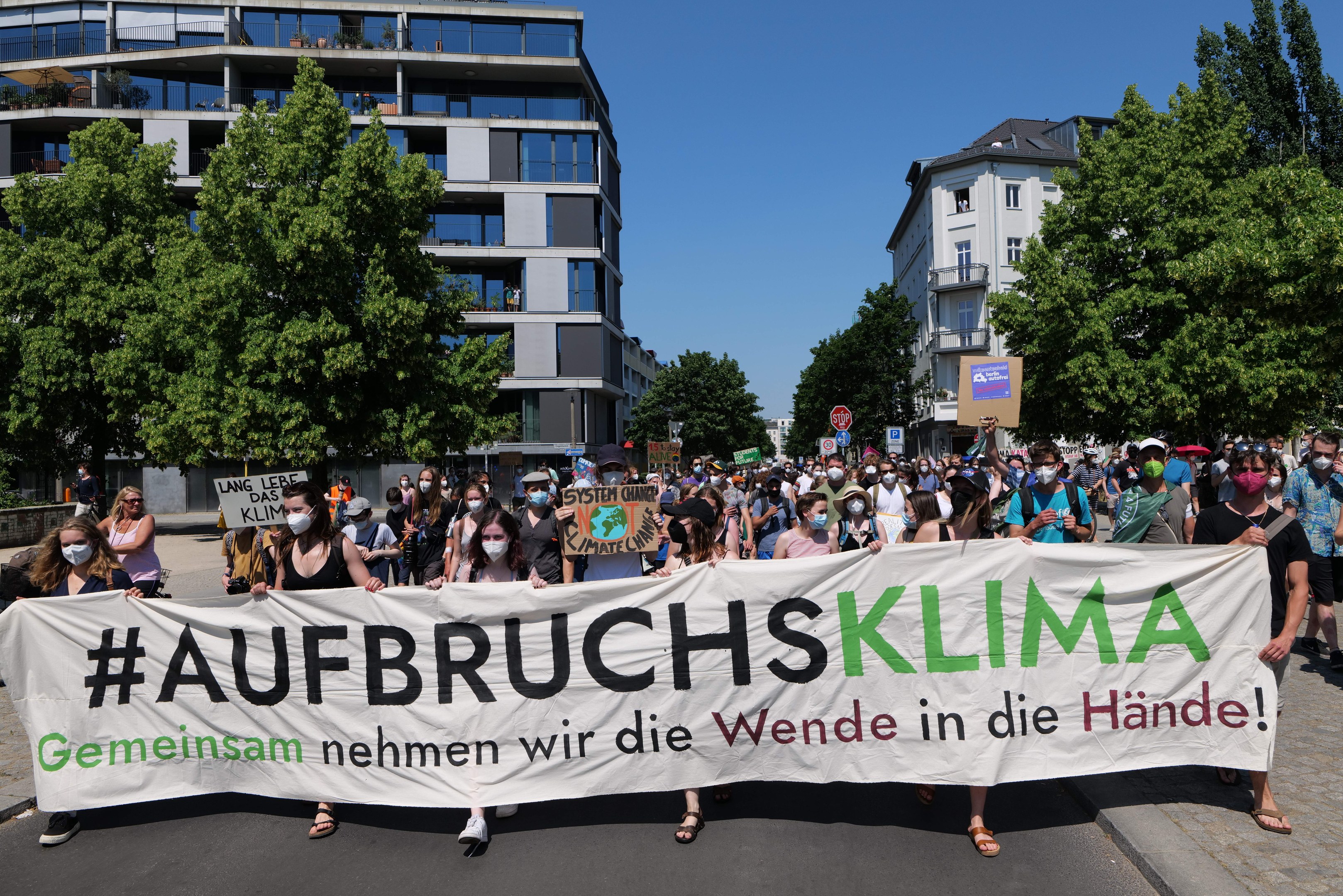 Eine Gruppe von Menschen mit Masken, die ein Banner mit der Aufschrift "Aufbruchsklima" vor einem Gebäude mit Bäumen und einem klaren Himmel halten.