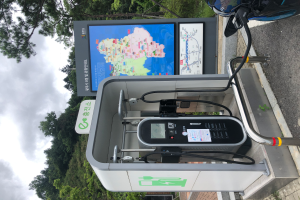 Eine Ladestation für Elektrofahrzeuge am Straßenrand mit einem Motorrad in der Nähe, Bäumen und einem klaren blauen Himmel im Hintergrund und einer digitalen Anzeigetafel, die anzeigt, dass es sich um eine E-Bike-Ladestation handelt.