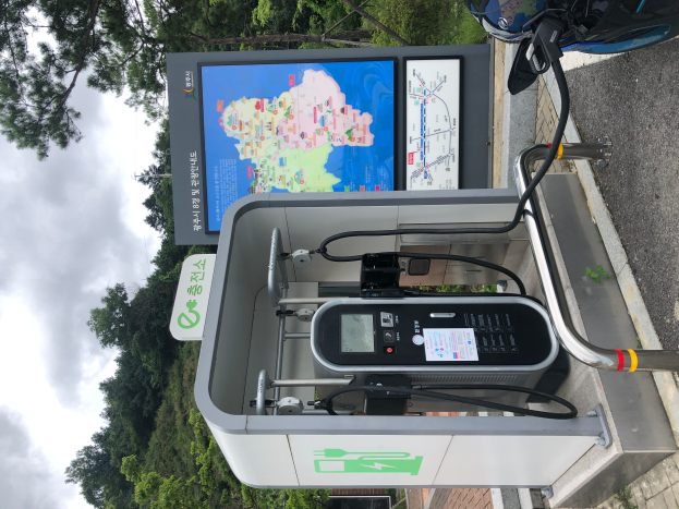 Eine Ladestation für Elektrofahrzeuge am Straßenrand mit einem Motorrad in der Nähe, Bäumen und einem klaren blauen Himmel im Hintergrund und einer digitalen Anzeigetafel, die anzeigt, dass es sich um eine E-Bike-Ladestation handelt.