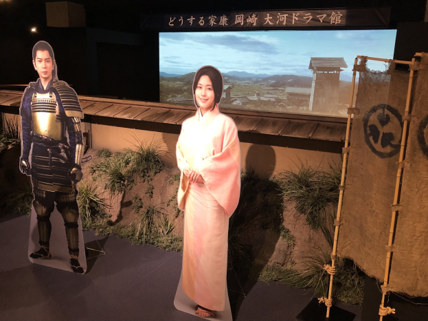 Zwei Menschen in traditioneller japanischer Kleidung - eine Frau in einem rosafarbenen Kimono und ein Mann in Samuraikleidung - lächeln gemeinsam auf einem Set mit Pflanzen, einem Bildschirm und Beleuchtungsausrüstung.