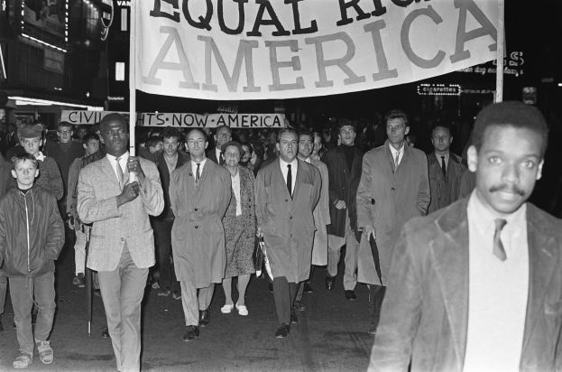Schwarz-weißes Foto einer Gruppe von Menschen, die eine Straße entlang marschieren und ein Banner mit der Aufschrift "Gleiche Rechte Amerika" tragen, mit Gebäuden und Straßenlaternen am Straßenrand.