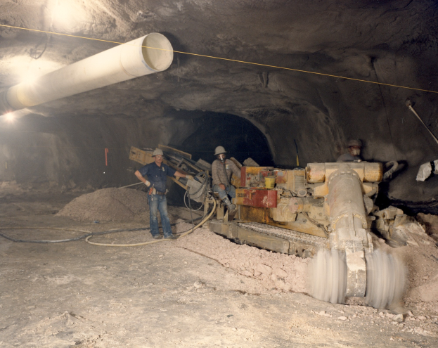 Gruppe von M├Ąnnern in Helmen bei der Arbeit in einem dunklen Stollensystem mit Maschinen und Rohren.