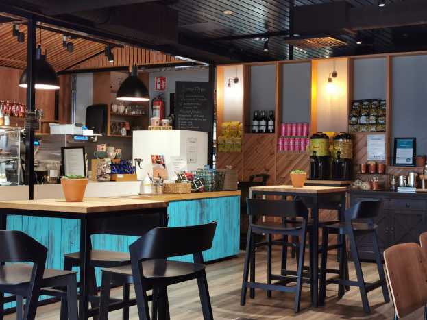 Innenansicht eines Restaurants mit Holztischen, Stühlen, einer Topfpflanze auf einem Tisch, einer Schiefertafel, einem Kühlschrank, einem Feuerlöscher, Regalen mit Flaschen, Schränken mit Gegenständen, einem gerahmten Foto an der Wand und Deckenlampen.