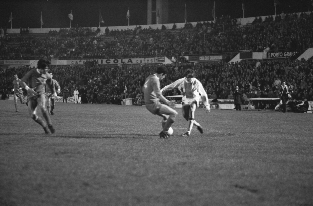 Schwarzes und weißes Foto von Männern, die Fußball in einem Stadion spielen, mit Zuschauern auf den Tribünen und Fahnen im Hintergrund.