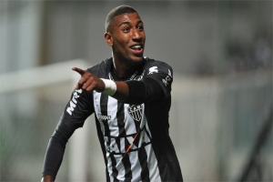 Fußballspieler in einem schwarz-weißen gestreiften Trikot, der lachend und ein Tor feiernd gezeigt wird, Hintergrund unscharf.