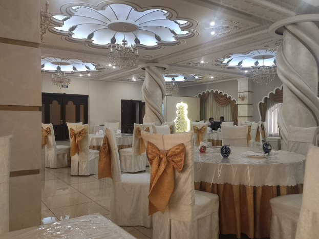 Großer Festsaal mit Tischen und Stühlen, mit Dekoration geschmückte Stühle, Wände mit Türen und Vorhängen geschmückt, Deckenleuchten und Kronleuchter schaffen eine gemütliche Atmosphäre.