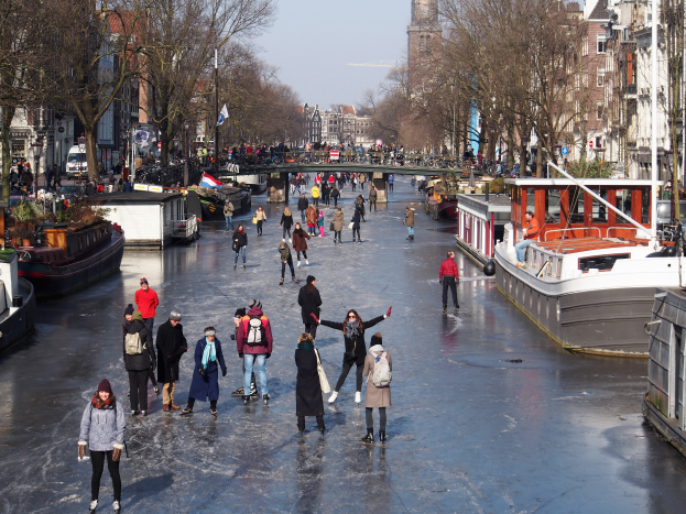 Eine Gruppe von Menschen, die auf einem zugefrorenen Kanal in Amsterdam Schlittschuh laufen, umgeben von Booten, Bäumen, Gebäuden, Laternenmasten, Flaggen und einer Brücke unter einem klaren Himmel.