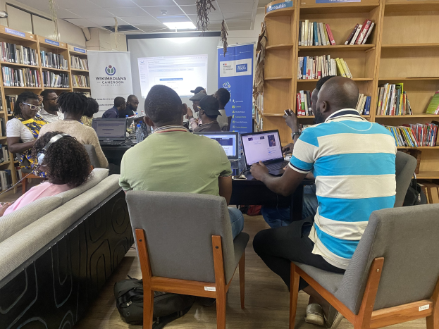 Gruppe von Menschen an einem Tisch mit Laptops umgeben von Bücherregalen, mit einem Banner, Bildschirm und Deckenbeleuchtung im Hintergrund und Taschen auf dem Boden.