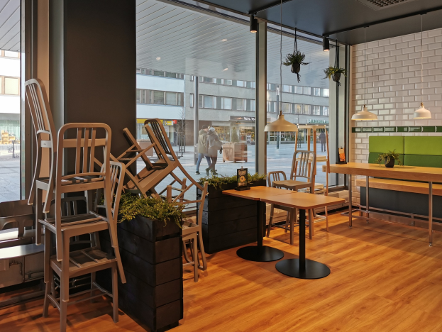Ein Restaurant-Innenraum mit Holztischen und -stühlen, große Fenster mit Blick auf eine Berliner Straße mit Passanten, Gebäuden und Straßenelementen.