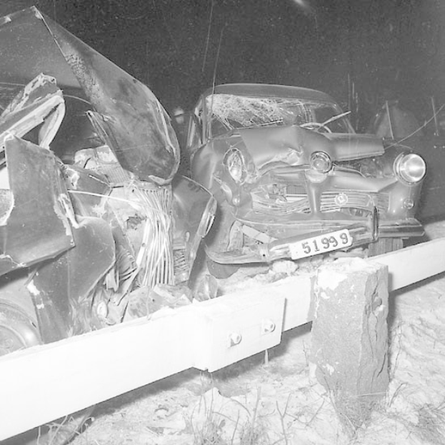 Schwarzes und weißes Foto eines beschädigten Autos mit eingedellter Motorhaube und zersplitterten Fenstern, das auf dem Straßenrand geparkt ist, umgeben von Gras und einem Zaun.