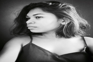 Eine selbstbewusste Frau in einem schwarzen Kleid posiert in einem Schwarz-Weiß-Foto, ihr glatter Bob und das dezente Make-up unterstreichen ihre selbstbewusste Haltung an der Wand.